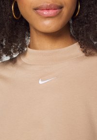 Detailní záběr na osobu s zlatými kruhovými náušnicemi a béžovým topem se malým bílým logem Nike na hrudi.
