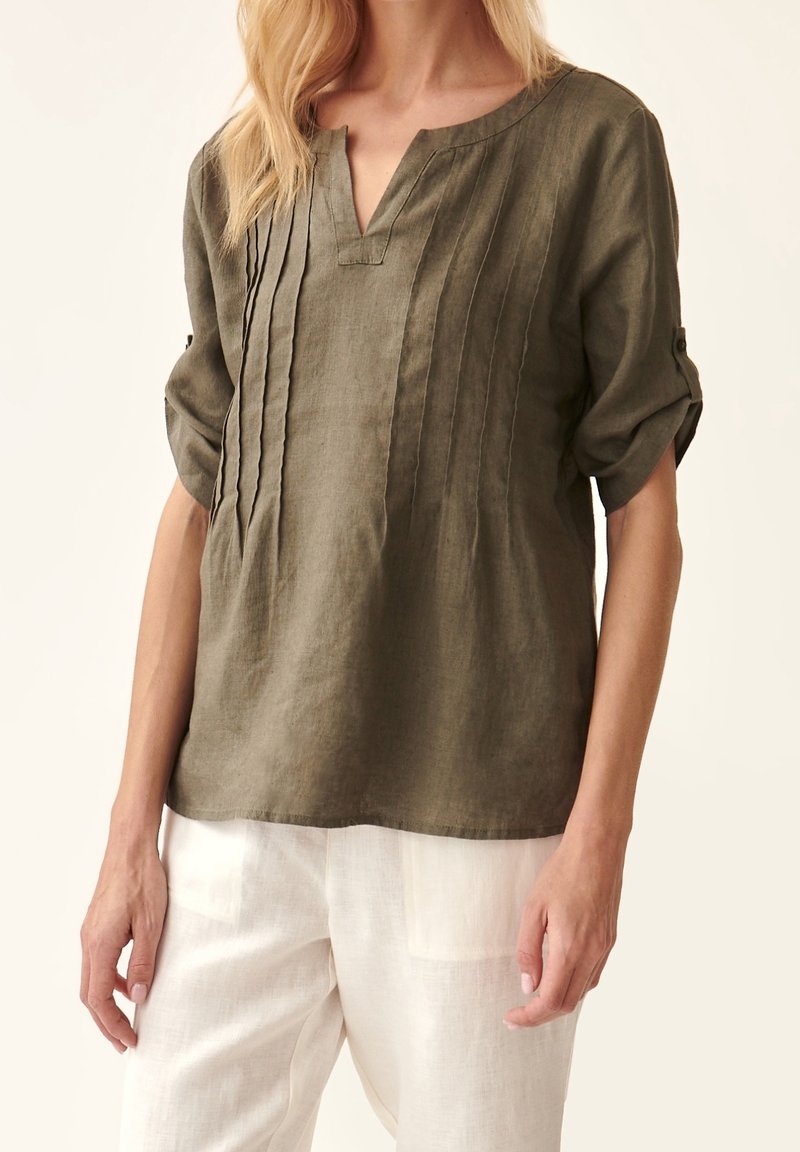 Femme portant un chemisier en lin vert olive avec des plis nervurés et des manches retroussées, associé à un pantalon en lin blanc.