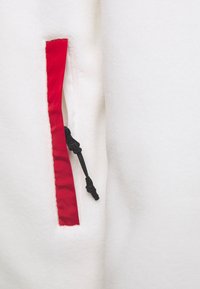Weißes Fleece-Oberteil mit einer roten Reißverschlusstasche und schwarzem Kordelzug. Die Textur wirkt weich und warm.