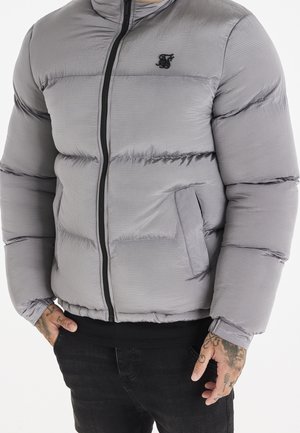 Homme portant une doudoune gris clair avec fermeture éclair noire et logo, mains dans les poches, pantalon noir, tatouages sur les mains, fond neutre.