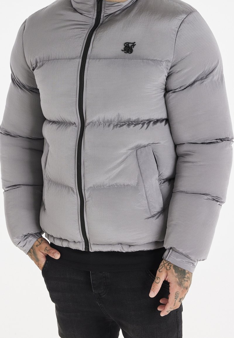 Homme portant une doudoune gris clair avec fermeture éclair noire et logo, mains dans les poches, pantalon noir, tatouages sur les mains, fond neutre.