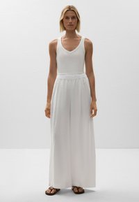 Top bianco senza maniche con scollatura a barca abbinato a pantaloni bianchi a gamba larga con cinturino elastico. Infradito marroni completano il look.