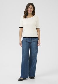 Haut à manches courtes blanc côtelé avec des accents noirs, associé à un jean large en denim bleu et des ballerines noires, se tenant sur un fond uni.