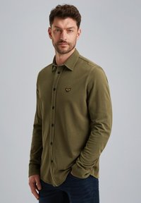 Camisa de manga larga verde oliva con botones, confeccionada en un tejido suave. Presenta un cuello, botones negros y un pequeño logo en el lado izquierdo del pecho.