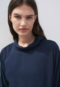 Mujer joven con cabello largo y castaño, vestida con una blusa azul oscura, mira con confianza a la cámara contra un fondo de color claro.