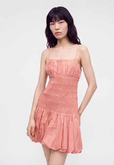 Jeune femme aux longs cheveux foncés portant une robe rose sans manches froncée avec une jupe plissée, debout devant un fond clair uni.