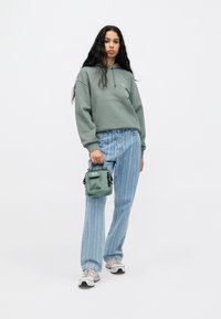 Groene hoodie met een voorzak, lichtblauwe gestreepte jeans, grijze sneakers en een kleine groene tas met een rits en logo-detail.