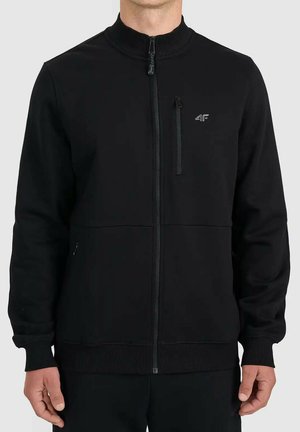 Mężczyzna noszący czarną kurtkę z pełnym zamkiem błyskawicznym, zapinaną kieszenią na piersi oraz ściągaczami przy mankietach i u dołu, stojący na jednolitym tle.