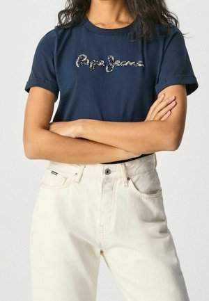 Mujer con camiseta azul marino con el logo de "Pepe Jeans" en cuentas y vaqueros blancos de talle alto, con los brazos cruzados sobre el pecho.