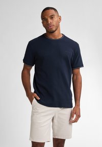 Námorně modré tričko s krátkým rukávem má strukturovanou látku a kontrastní světle barevné švy. Kombinováno se světlými béžovými šortkami.