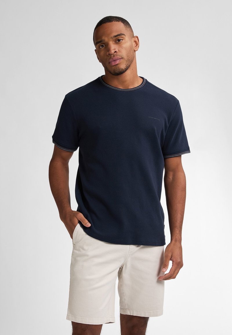 Námorně modré tričko s krátkým rukávem má strukturovanou látku a kontrastní světle barevné švy. Kombinováno se světlými béžovými šortkami.