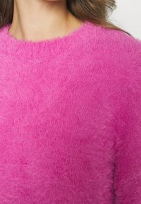 Luddigt rosa tröja med rundad halsringning. Har mjukt, texturerat material och en löst sittande design som betonar värme och komfort.