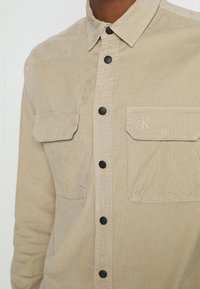 Chemise beige boutonnée avec deux poches poitrine à rabat, boutons noirs et logo brodé "cK" sur la poche droite.
