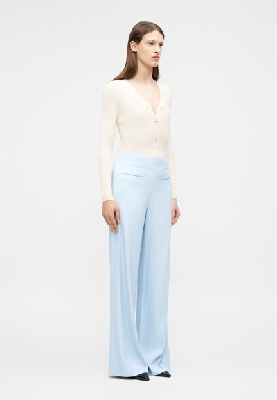 Γυναίκα που στέκεται φορώντας μια κρεμ ζακέτα με V-λαιμόκοψη και κουμπιά, καθώς και ανοιχτό μπλε παντελόνι φαρδιάς γραμμής με μαύρα μυτερά παπούτσια.