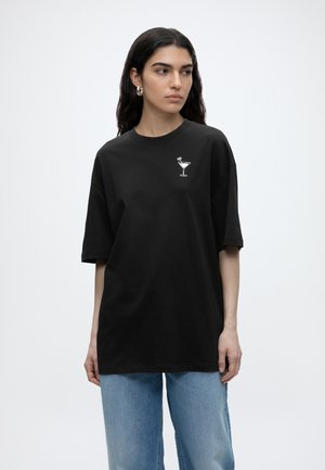 Femme aux longs cheveux foncés portant des boucles d'oreilles argentées en anneau, un t-shirt noir ample avec un motif de verre à cocktail, et un jean bleu clair, regardant sur le côté.