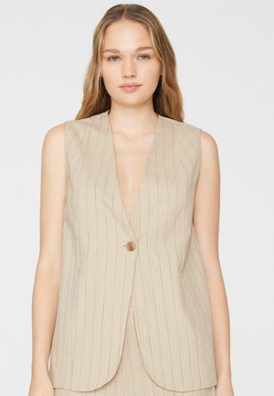 Jeune femme aux longs cheveux blonds portant un blazer sans manches beige à fines rayures avec un seul bouton sur fond blanc.