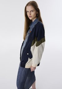 Marineblauwe en roomkleurige windbreaker jas met een denim kraag, voorzien van een elastische tailleband, zijzakken en contrasterende groene strookdetails.