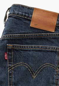 Jean en denim bleu foncé de Levi's avec une patte en cuir marron, des coutures jaunes et un design arqué emblématique sur la poche arrière. Étiquette rouge attachée.