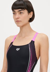 Μαύρο μαγιό με ροζ τιράντες και λευκές λεπτομέρειες. Διαθέτει διαγώνιες ροζ γραμμές. Λογότυπο στο στήθος. Ομαλό, εφαρμοστό υλικό.