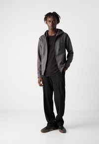 Felpa grigia con zip e cappuccio, maglietta nera e pantaloni neri larghi. L'outfit presenta un design casual, tessuto testurizzato e accenti sottili.