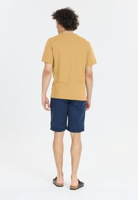 En tanfärgad bomullströja med korta ärmar, matchad med mörkblå shorts. Sandaler fulländar outfiten. Utsikten visar baksidan.