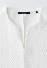 Top bianco con texture, scollatura a V e un sottile motivo geometrico. Presenta un'etichetta con la scritta "someday" e una tag di taglia "36".