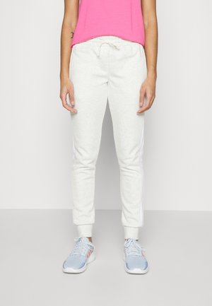 Person trägt weiße Jogginghosen mit Kordelzug, hellblaue und pinke Turnschuhe sowie ein leuchtend pinkes ärmelloses Oberteil und steht vor einem schlichten Hintergrund.