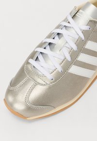 Tenis prateado com um upper metálico texturizado, cordões brancos e três riscas brancas ao longo do lado. Bico arredondado suave e sola de borracha.