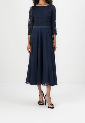 Femme portant une robe bleu marine à manches trois-quarts en dentelle et jupe fluide, assortie à des sandales noires à talons hauts avec des lanières argentées.