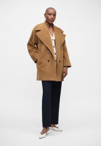 Καφέ πανωφόρι oversized από μίγμα μάλλινου υλικού, με μεγάλες κολάρα και διπλές κουμπότρυπες, σε συνδυασμό με λευκή πουκάμισο και μπλε παντελόνι. Λευκά παπούτσια.