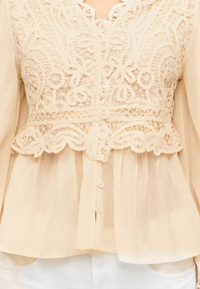 Μπεζ μπλούζα με περίτεχνη δαντέλα στο πάνω μέρος του στήθους και κουμπιά στο κέντρο, φορεμένη πάνω από λευκό παντελόνι.