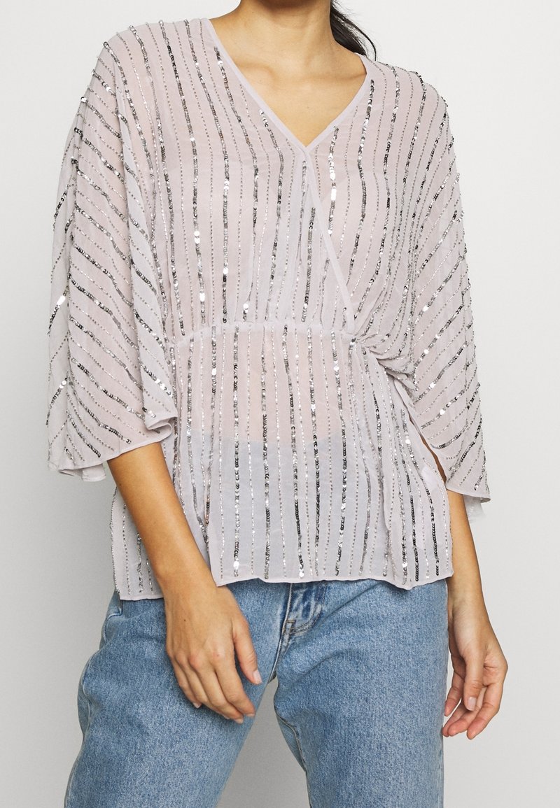 Femme portant un chemisier rose clair à col en V avec des rayures verticales en sequins argentés et des manches larges, associé à un jean bleu.