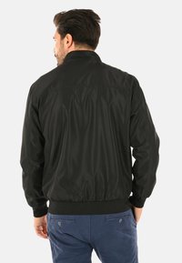 Giubbotto bomber nero con una texture liscia e lucida, polsini e orlo a coste, e un design minimalista. Vista posteriore che mette in risalto una silhouette semplice e aderente.