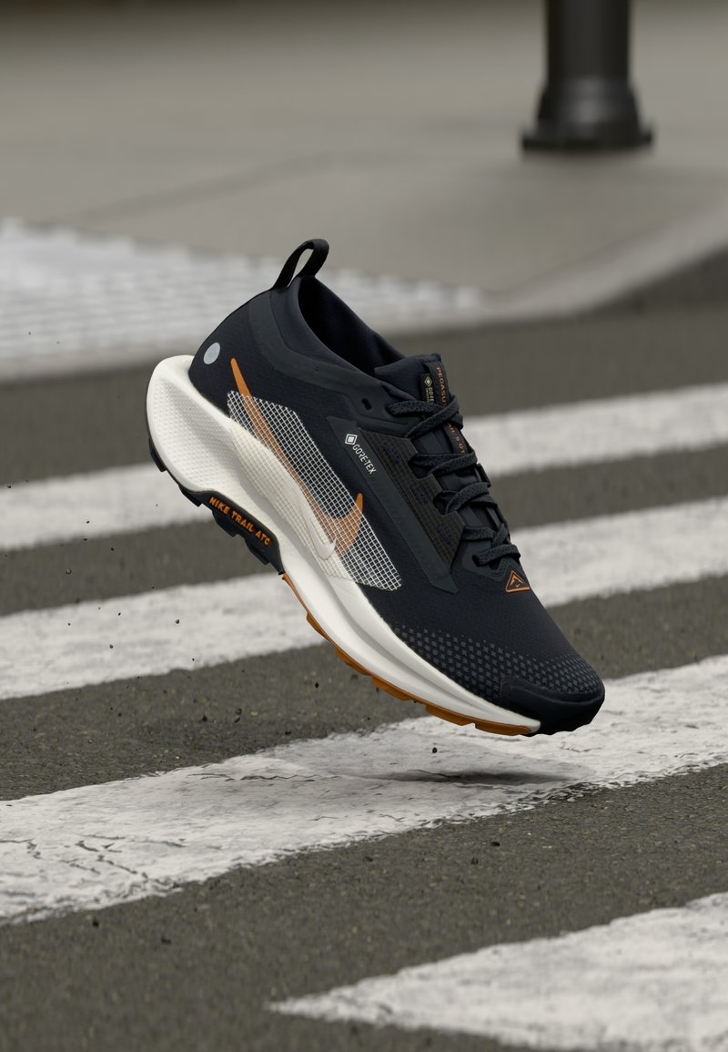 Chaussure de trail Nike en noir et blanc avec des accents orange flottant au-dessus d'un passage piéton zébré sur une route en asphalte.