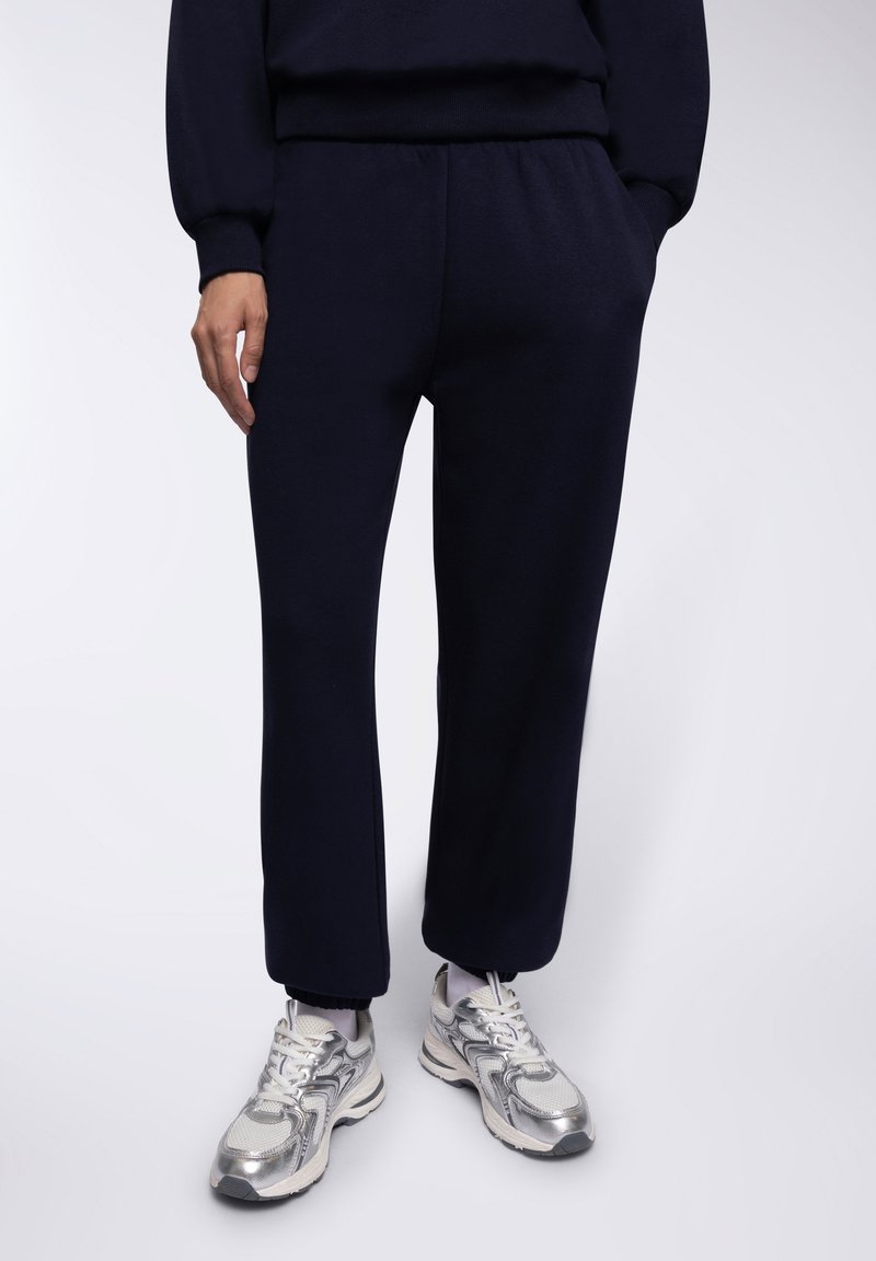 Granatowe dresy wykonane z miękkiego materiału, z elastycznym pasem i prostym krojem, zestawione z metalicznie srebrnymi butami sportowymi.