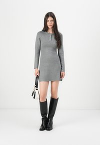 Robe grise à manches longues avec un col boutonné et des fentes latérales, associée à des bottes noires montantes. Texture lisse, design ajusté.