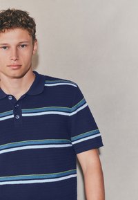Tricou polo bleumarin cu țesătură texturată, cu dungi orizontale în albastru deschis și verde. Guler cu două nasturi și mâneci scurte.