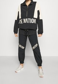 Svart och krämfärgad vindjacka med halvdragkedja, elastiska ärmmuddar och texten "RE NATION." Matchande svarta joggers med samma text.