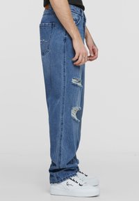 Jeans in denim azzurro chiaro con vestibilità comoda, caratterizzati da toppe strappate e bordi grezzi, indossati con sneakers bianche.