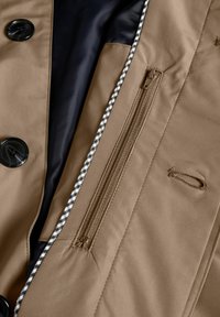 Brown outerwear featuring a dark navy lining, large black buttons, and a zippered pocket with a checkered accent trim. Smooth texture.