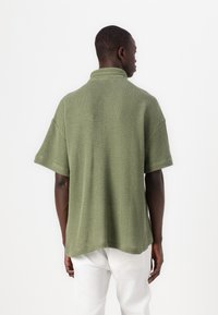 Maglione polo lavorato a maglia verde con vestibilità rilassata, maniche corte e finitura testurizzata. La vista posteriore mostra un colletto e una silhouette morbida.