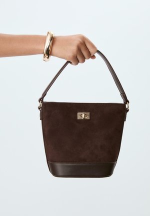 Hand, die eine dunkelbraune Wildleder- und Leder-Bucket-Handtasche mit goldenem Verschluss und passendem Armband am Handgelenk hält.
