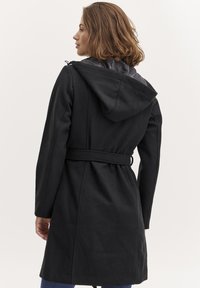 Cappotto in misto lana nera con vita cintrata, cappuccio e fodera interna liscia. Mostra la vista posteriore, evidenziando le cuciture e gli accenti della cintura.