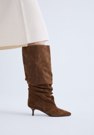Bota marrón de ante blanda hasta la rodilla con punta afilada y tacón bajo estilo kitten, combinada con una falda beige clara sobre un fondo liso.