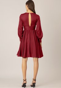 Burgundy kort klänning med en figursydd midja, puffärmar och en nyckelhål på baksidan. Tillverkad av ett mjukt tyg med en flödande kjoldesign.