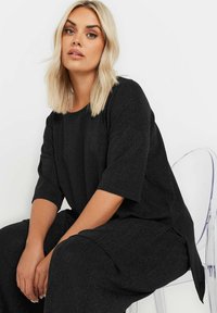 Top in maglia nera a costine con maniche corte e vestibilità rilassata. Presenta spacchi laterali e una leggera lucentezza. Modella seduta su una sedia trasparente.