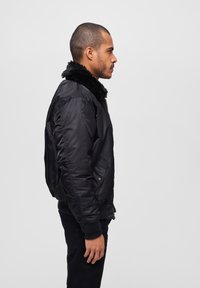 Chaqueta bombers negra con un cuello de peluche, puños acanalados y cierre de cremallera. Incluye bolsillos laterales y una textura de tela suave y brillante.