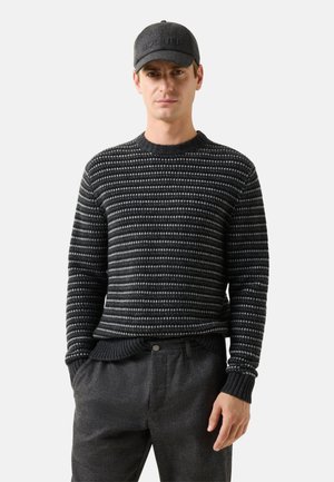 Man draagt een zwart-grijs gestreepte gebreide trui, donkergrijze broek en een zwarte baseballpet, staat met handen in de zakken.
