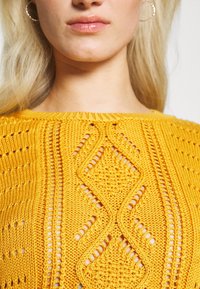 Gul stickad tröja med öppna mönster och en texturerad design, med rund halsringning och synlig stickdetalj.