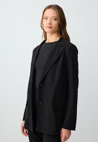 Blazer nero con una vestibilità sartoriale, revers a notched, due bottoni e tasche laterali. Tessuto liscio e leggero. Indossato sopra un top impreziosito.